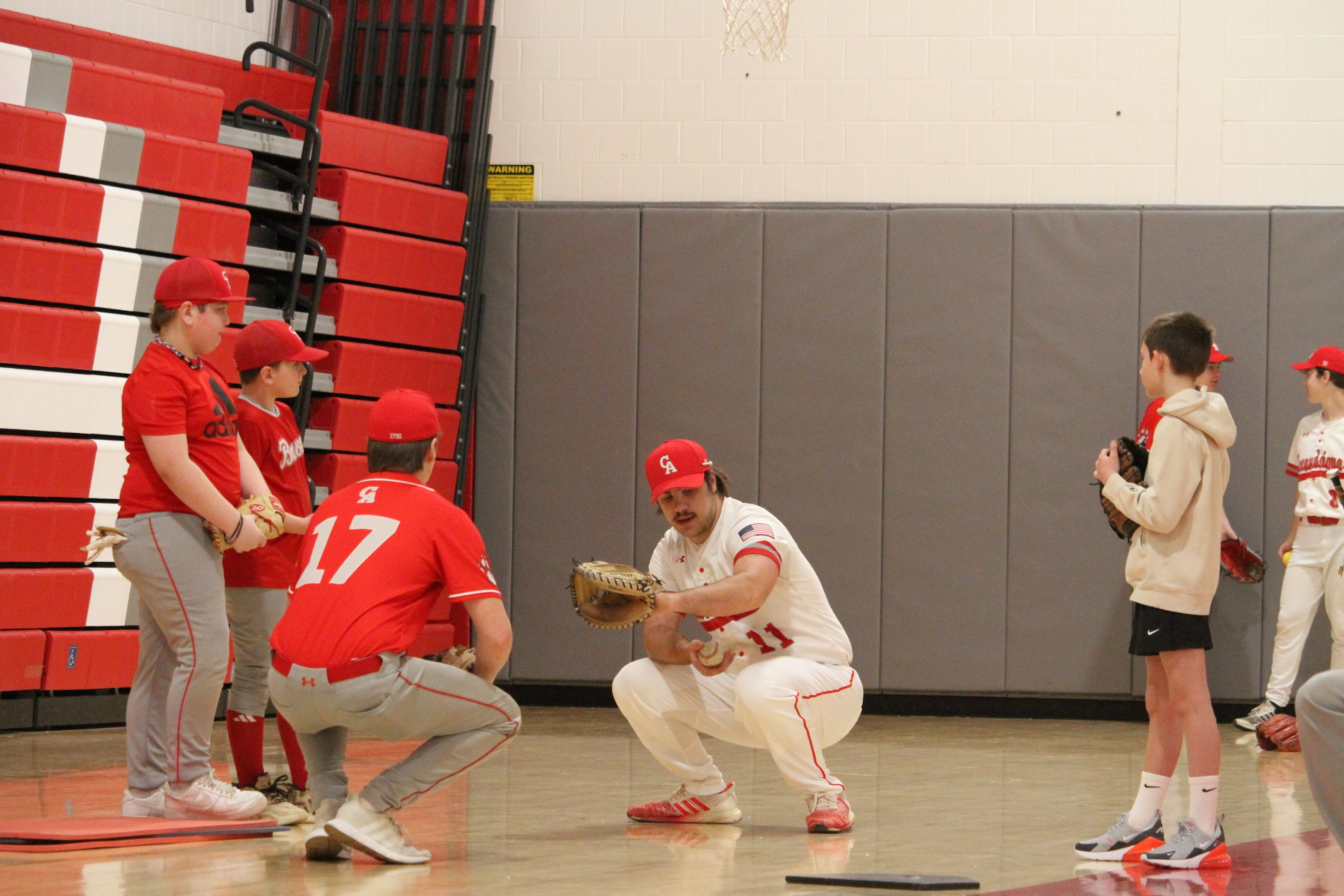 A baseball player crouches down to demonstrate how to catch a pitch while younger athletes look on.
